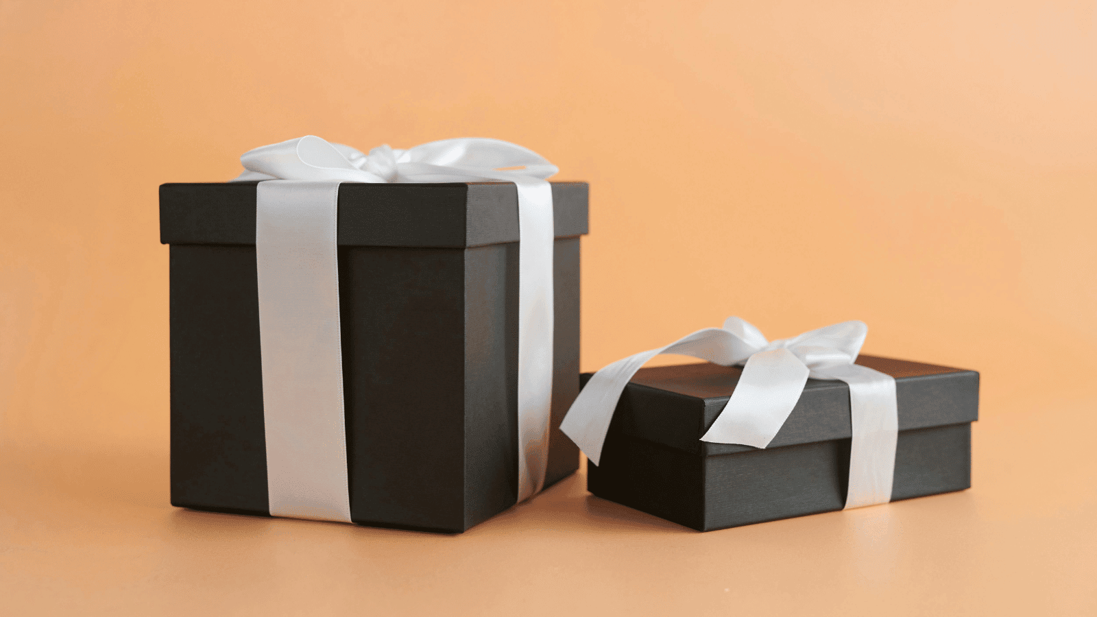 Two black gift boxes of different sizes resting on a peach-colored background. Each box is elegantly wrapped with a wide white satin ribbon tied in a bow.