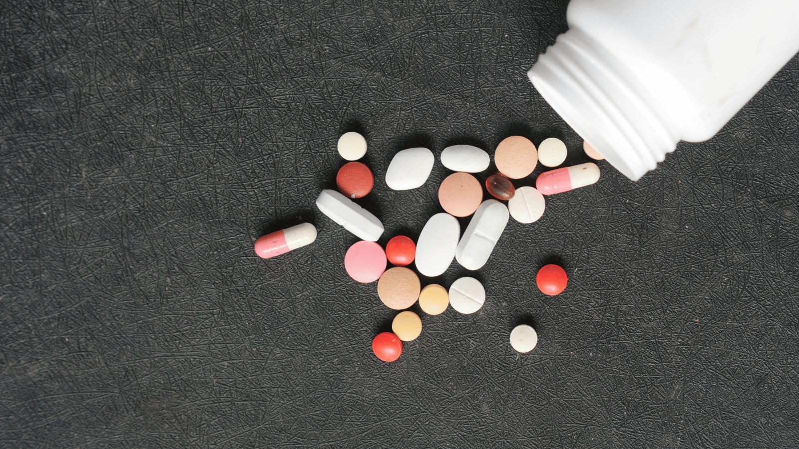 A high-angle, close-up shot of various medical pills and capsules scattered across a dark, textured gray surface. A white plastic medicine bottle is tipped over in the upper right corner, with colorful medication in shades of red, white, pink, and tan spilling out.